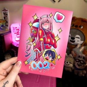 May include: A rectangular art print on a pink background featuring an anime character with long pink hair in a red suit, a smaller character with dark hair, the text 002, Japanese characters, a heart icon, and various yellow star accents.