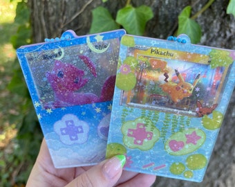 Könnte beinhalten: Zwei durchsichtige Schlüsselanhänger aus Kunststoff mit blauem und weißem Hintergrund. Die Schlüsselanhänger zeigen ein Pokémon-Kartendesign mit einer rosa und weißen Pokémon-Karte links und einer gelben und weißen Pokémon-Karte rechts. Der Text auf den Karten lautet "Pikachu" und "Mewtwo".