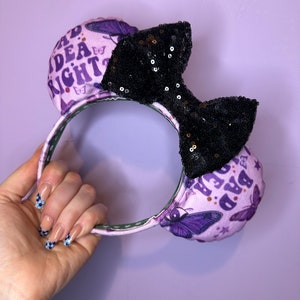 May include: Purple and black mouse ears with a black sequin bow. The ears are decorated with butterflies and the text "Bad Idea Right?" and "Bad Idea Right?"