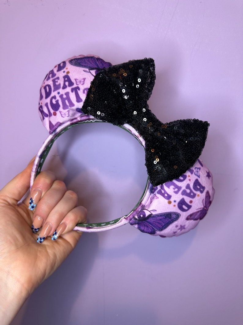May include: Purple and black mouse ears with a black sequin bow. The ears are decorated with butterflies and the text "Bad Idea Right?" and "Bad Idea Right?"