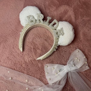 May include: A white bridal headband with the word "BRIDE" in sparkling letters. The headband is decorated with pearls and has two sequined ears. A white veil with pearl accents is also visible.