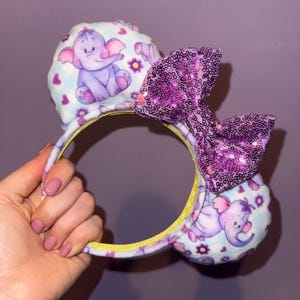 May include: Mouse ear headband with a purple sequined bow and plush ears. The ears have a light blue background with purple elephants, hearts, and flowers. The headband is yellow.