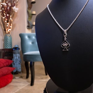 May include: A silver chain necklace with a black spade pendant featuring a crown with clear stones. The necklace is displayed on a black velvet stand.