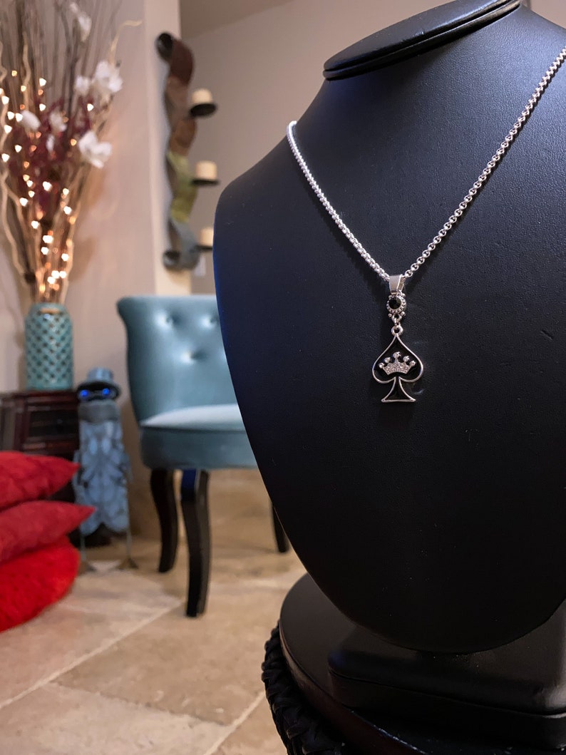 May include: A silver chain necklace with a black spade pendant featuring a crown with clear stones. The necklace is displayed on a black velvet stand.