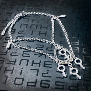 Puede incluir: Un collar de cadena de plata con un colgante en forma de l&aacute;grima que presenta un s&iacute;mbolo femenino estilizado y tres colgantes m&aacute;s peque&ntilde;os con s&iacute;mbolos masculinos estilizados.