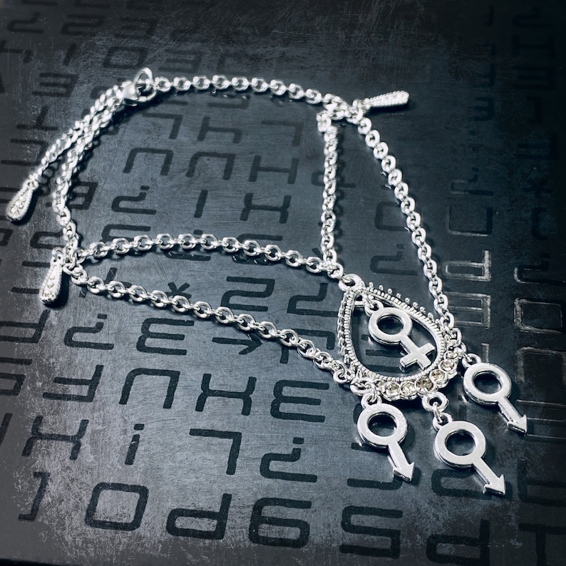 Puede incluir: Un collar de cadena de plata con un colgante en forma de l&aacute;grima que presenta un s&iacute;mbolo femenino estilizado y tres colgantes m&aacute;s peque&ntilde;os con s&iacute;mbolos masculinos estilizados.
