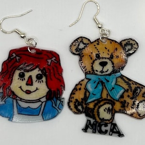 May include: A pair of earrings featuring a hand-painted rag doll and a teddy bear. The rag doll has red hair, a blue dress, and a white face. The teddy bear is brown with a blue bow tie. Both earrings are made of clear plastic and have silver hooks.