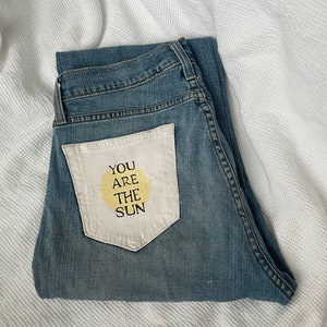 May include: Blue denim jeans with a white patch pocket on the back. The patch pocket has a yellow circle and the text "YOU ARE THE SUN" in black.