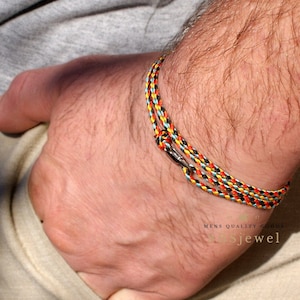 以下が含まれることがあります： 銀色の留め具が付いた、マルチカラーの編み込みブレスレット。ブレスレットは、黒、赤、黄、青の糸でできています。