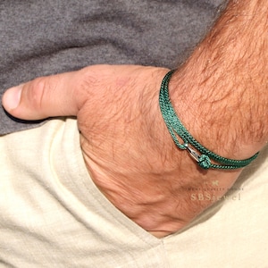 Può includere: Un braccialetto verde intrecciato con chiusura argentata. Il braccialetto è realizzato in un materiale sottile e intrecciato ed è assicurato con una piccola chiusura color argento. Il braccialetto è indossato al polso.