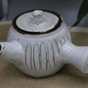 May include: White ceramic teapot with a lid and two spouts. The teapot has a rounded body with vertical grooves and brown accents. The lid has a small knob. The teapot is on a tan surface.