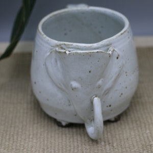 May include: White ceramic mug with an elephant design. The mug has a textured surface and a handle shaped like an elephant's trunk.