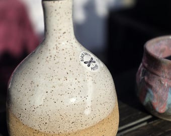 May include: A ceramic oil cruet with a speckled white glaze on the upper portion and a natural tan clay base. The bottle features a metal pour spout and a circular sticker with a dragonfly graphic and the text Black Earth Pottery.