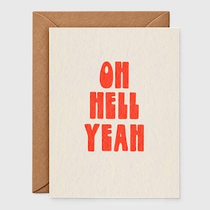 May include: A cream-colored greeting card with the text OH HELL YEAH printed in bold, textured red capital letters. The card is positioned in front of a brown kraft paper envelope, creating a simple contrast between the light and dark paper.