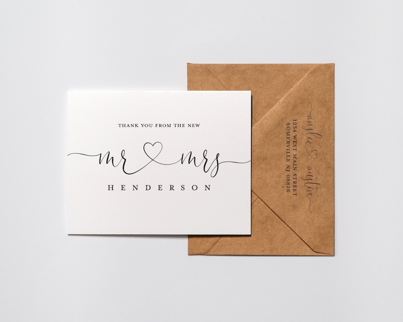 May include: A white thank you card with the text "Thank you from the new Mr & Mrs Henderson" in black script font with a heart symbol between the words "Mr" and "Mrs". The card is sitting on top of a brown envelope with the text "anglee & austin 1234 West Main Street Somerville NJ 08876" in black script font.