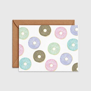 May include: A rectangular greeting card featuring a repeating pattern of donuts with pastel-colored frosting and sprinkles, including shades of pink, blue, green, and brown, paired with a plain brown kraft paper envelope positioned behind the card.