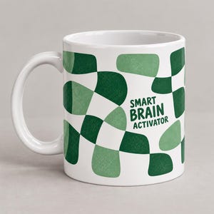 Peut inclure: Un mug en céramique blanche orné d'un motif à damier ondulé répétitif dans des tons de vert foncé et clair. Le texte SMART BRAIN ACTIVATOR est imprimé en lettres capitales vert foncé sur le côté du mug, intégré au motif.