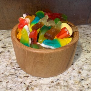 May include: A wooden bowl filled with colorful gummy candies, including fish, bottles, and worms. The bowl is sitting on a light brown countertop.