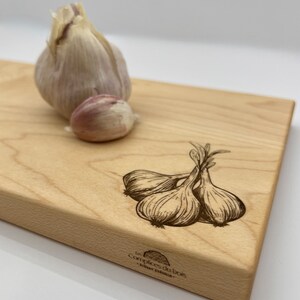 Puede incluir: Una tabla de cortar de madera con un dibujo de dientes de ajo y el texto "Les Complices du Bois - Artisan charcutier -"
