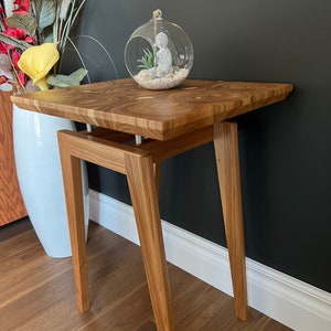May include: A small wooden end table with a light brown wood grain finish. The table has a rectangular top and four slender legs. A glass globe with a small white figurine inside sits on the table top.
