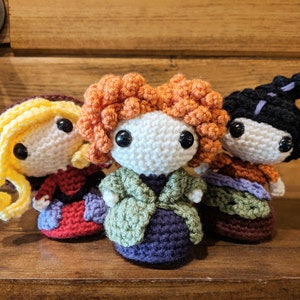 May include: Three crocheted dolls with different coloured hair and clothing. The doll on the left has yellow hair and a red and purple dress. The doll in the middle has orange hair and a green and purple dress. The doll on the right has black hair and a brown and purple dress.