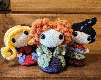 May include: Three crocheted dolls with different coloured hair and clothing. The doll on the left has yellow hair and a red and purple dress. The doll in the middle has orange hair and a green and purple dress. The doll on the right has black hair and a brown and purple dress.