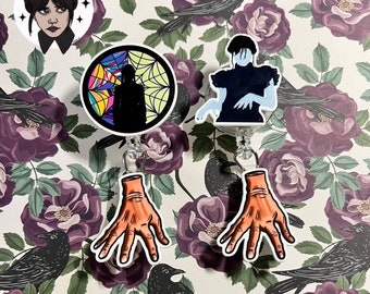 May include: Two badge reels featuring character designs. One shows a silhouette against a colorful spiderweb, the other a figure in a black dress. Both have a dangling orange hand charm attached to the bottom with a clear plastic strap.