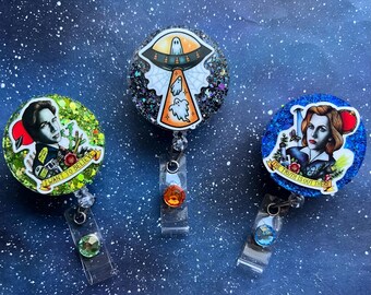 May include: Three round retractable badge holders with glittery backgrounds. One features a portrait and the text "I WANT TO BELIEVE", another has a UFO and ghosts, and the last has a portrait and the text "THE TRUTH IS OUT THERE".