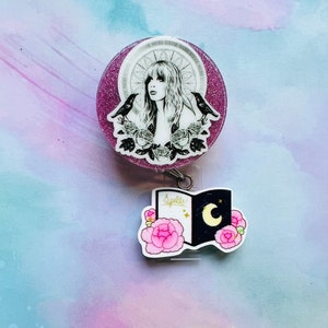 May include: A pink glitter badge reel with a black and white image of a woman with long hair, two black birds, and flowers. The badge reel has a black and white charm attached to it that says "Spells!" and has a crescent moon and stars on it.