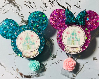 May include: Two glitter Mickey Mouse-shaped snow globe keychains. One is teal blue with silver glitter and the other is pink with silver glitter. Both have a white snow globe with a drawing of Cinderella's castle inside.