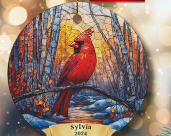 May include: A round ceramic ornament with a stained glass design of a cardinal bird in a winter forest scene. The ornament has a gold ribbon and a personalized name tag that reads "Sylvia 2024".