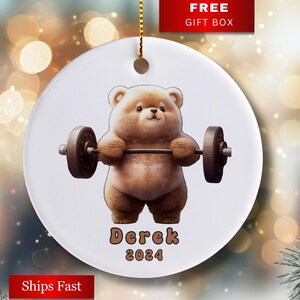 May include: A white ceramic ornament with a brown bear lifting a barbell. The bear is wearing a brown fur coat and has a white muzzle. The ornament has the name "Derek" and the year "2024" printed on it.