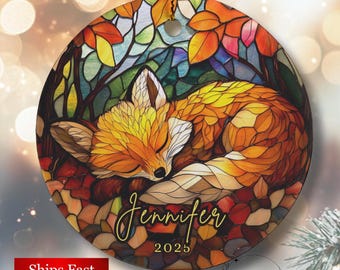May include: Round ornament featuring a sleeping fox in autumn colors. The fox is orange and brown, surrounded by colorful leaves. The name "Jennifer" and the year "2025" are written on the ornament. The ornament has a gold string for hanging.