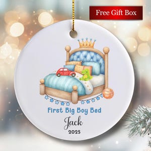 May include: A white ceramic ornament with a watercolor illustration of a bed, a red car, and a green dinosaur. The text "First Big Boy Bed" and the name "Jack" are printed on the ornament. A gold string is attached for hanging.