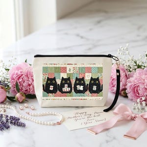 May include: A cream-colored zippered pouch featuring a graphic of four black cats sitting in front of a colorful patchwork quilt pattern. Each cat holds a small white tile. A black wrist strap is attached to the side of the pouch.