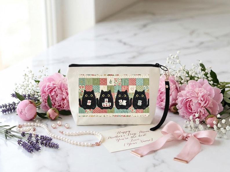 May include: A cream-colored zippered pouch featuring a graphic of four black cats sitting in front of a colorful patchwork quilt pattern. Each cat holds a small white tile. A black wrist strap is attached to the side of the pouch.