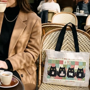 May include: A white canvas tote bag featuring a rectangular graphic print of four black cats sitting in a row against a colorful patchwork background. The cats are depicted with large yellow eyes and each holds a small white tile with red symbols.