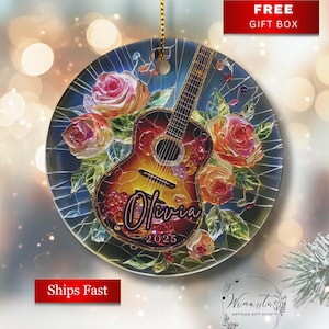 May include: Round ceramic ornament featuring a guitar design with roses and the name "Olivia" and the year "2025". The ornament has a gold string for hanging and a "Free Gift Box" label.