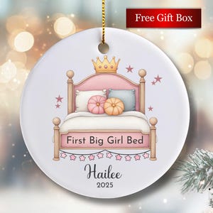 May include: A round, white ceramic ornament with a gold hanging loop. The ornament features a pink bed with a gold crown, pillows, and the text "First Big Girl Bed." Below is the name "Hailee" and the year "2025."