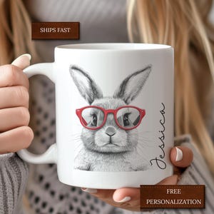 May include: White ceramic mug with a black and white illustration of a bunny wearing red glasses. The mug has the name "Jessica" written on the side.