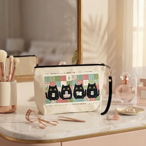 May include: A cream-colored canvas zippered pouch featuring a graphic print of four black cats sitting in a row against a colorful patchwork quilt background. The pouch includes a black wrist strap attached to the side zipper pull.