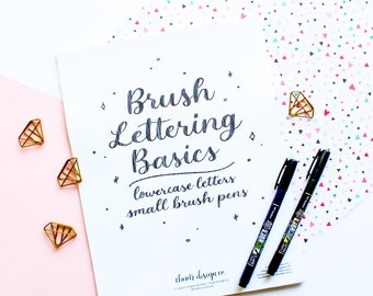 May include: A white sheet of paper with the words "Brush Lettering Basics" written in black cursive. Below the title, it says "lowercase letters" and "small brush pens". The paper is on a white surface with a pink surface to the left and a white surface with a pink and blue pattern to the right. Two black brush pens are on the patterned surface.
