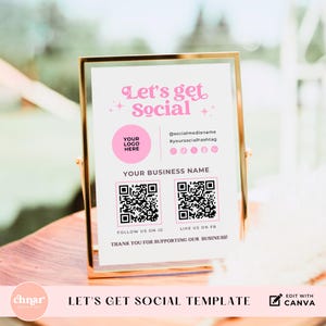 Op de afbeelding: Een wit bord met een roze rand en de tekst "Let's get Social" in roze. Het bord bevat ook een ruimte voor een logo, social media-handvatten en twee QR-codes. De tekst "Follow us on IG" en "Like us on FB" staat onder de QR-codes. Het bord staat op een houten tafel.