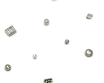 May include: Assortment of silver-toned metal beads for jewelry making. The beads include a heart, a circle, a square, a triangle, a cylinder, and a few round beads.