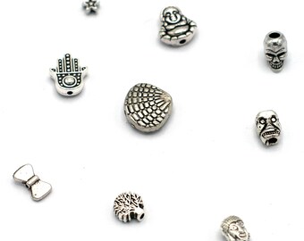 May include: Assortment of silver charm beads, including a skull, a hand, a tree, a bow tie, a Buddha, and a scale pattern.