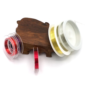 Op de afbeelding: Vier spoelen draad voor het maken van sieraden. De spoelen zijn van helder plastic en bevatten rood, goud, zilver en paars draad. De spoelen liggen op een houten basis.