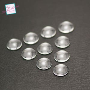 Op de afbeelding: Een groep van 10 heldere glazen koepelvormige cabochons. De cabochons zijn in een halve cirkel op een donkergrijs oppervlak gerangschikt.