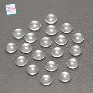 Op de afbeelding: Een verzameling van 20 heldere glazen cabochons, elk ongeveer 2,5 cm in diameter. De cabochons zijn in een cirkelvormige opstelling op een grijs oppervlak geplaatst.