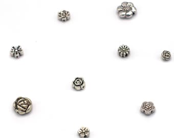 May include: Assortment of silver metal flower charms. The charms are in various shapes and sizes, including roses, daisies, and other floral designs.