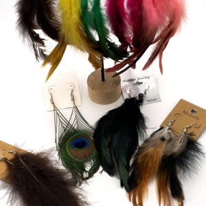 Peut inclure: Assortiment de boucles d'oreilles en plumes de différentes couleurs et styles, notamment des plumes de paon, noires, brunes et multicolores. Les boucles d'oreilles sont présentées sur une surface blanche avec des cartes qui disent "Fashion Jewelry" et "Michelle".